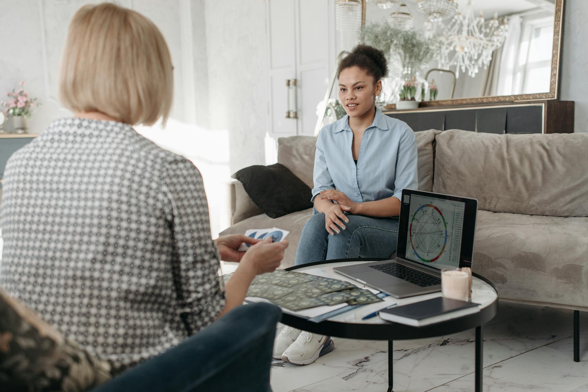 Women engaged in a tarot reading session with astrology charts on a laptop in a cozy living room.